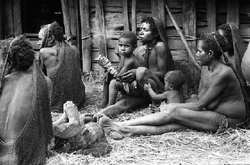 Children - one has fingers bandaged; sitting with a woman outside of hunu at Abulupak