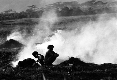 2 men sitting on the ground above the ditch in front of smoke that is coming up from trash piles behind them