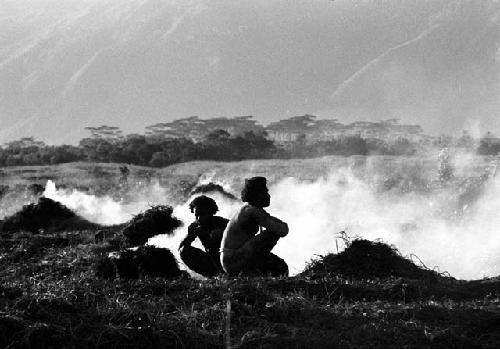 2 men sitting on the ground above the ditch in front of smoke that is coming up from trash piles behind them