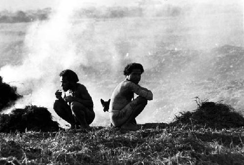 2 men sitting on the ground above the ditch in front of smoke that is coming up from trash piles behind them