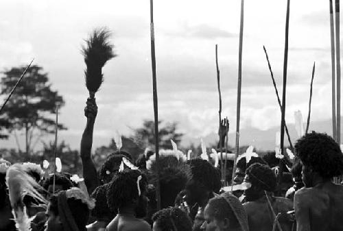 Group of warriors singing; one lifts a sué laré in the air