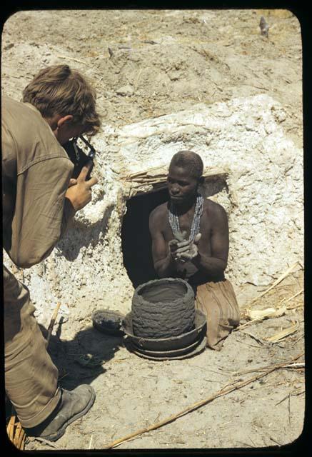 Woman sitting in front of a cave, making a pot, with John Marshall photographing her
