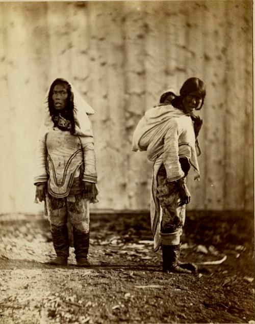 Two Inuit women wearing traditional dress, one carrying a baby on her back