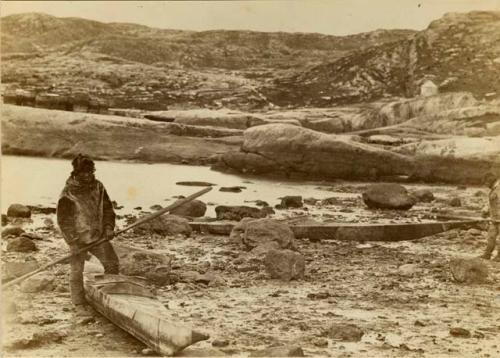 Two Inuit men with kayaks on rocky beach