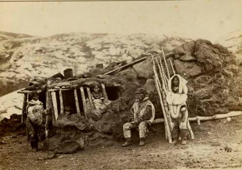 Inuit man, woman and child with two dogs in front of sod house