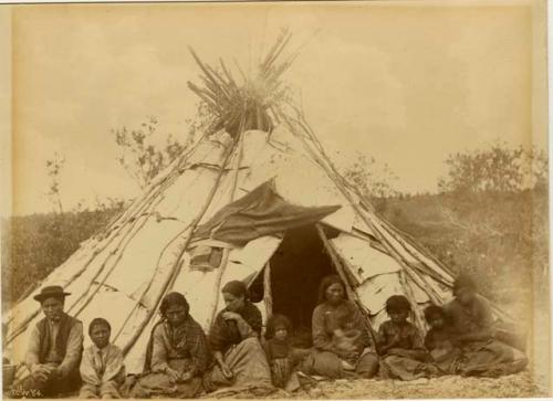 Cree man, four women, five children seated in front of birch bark covered tepee