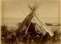Group of Cree Indians seated in front of birch bark covered tepee