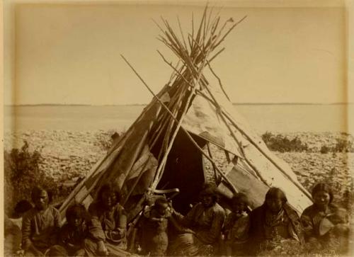 Group of Cree Indians seated in front of birch bark covered tepee