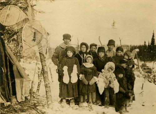 Three men, three(?) women, five children and a dog standing in snow