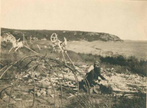 Two people seated near tent-bough frame of sweat lodge near lakeshore