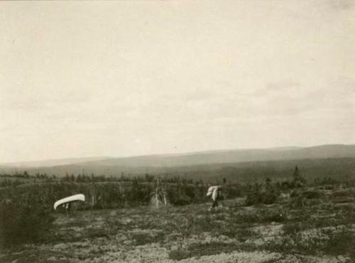 Two figures walking across meadow. One with burden on back and other with canoe