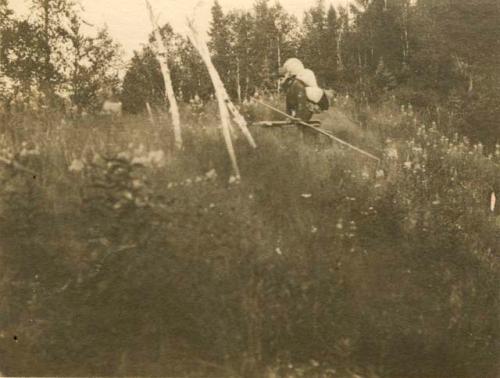 Man walking across meadow with burden on back and several poles in his hand.