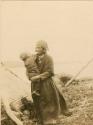 Naskapi woman holding a child and standing next to a lodge