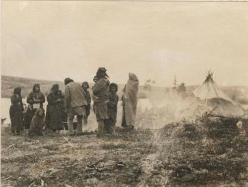 Group of men, women and children around a fire at camp by lake.