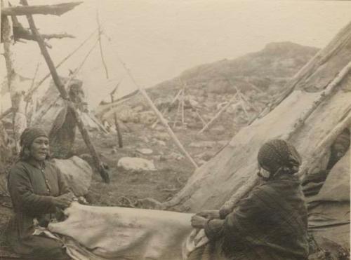 Two women working a skin. Covered tepee to right. Tepee frames in background.