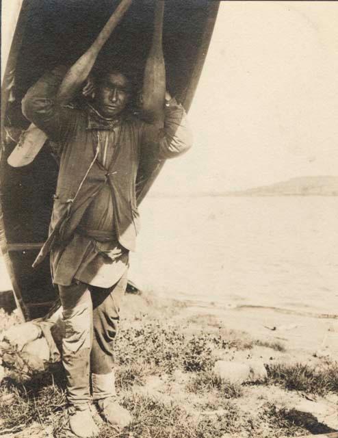 Man carrying canoe on shoulders - paddles lashed to make shoulder rests