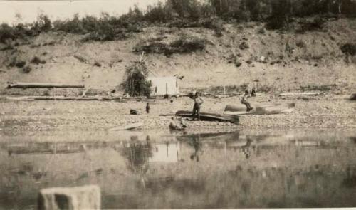 Two men near two canoes on river bank. Dwelling structure in background