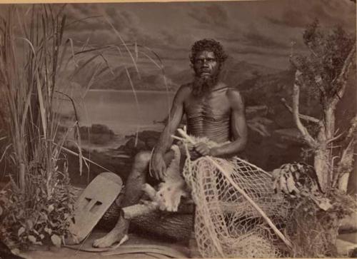 Studio-staged bush scene featuring an Aboriginal man of New South Wales, seated and gripping a fish net