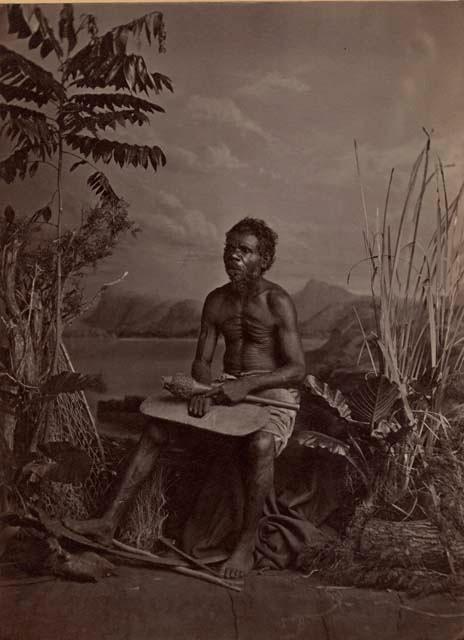 Studio-staged bush scene featuring an Aboriginal man of New South Wales, seated and holding a club