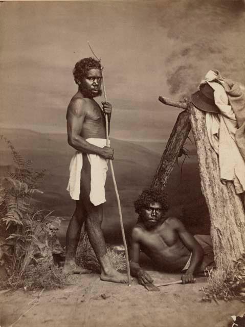 Studio-staged bush scene featuring two Aboriginal men, one standing holding a long stick, the other lying beneath a wooden shelter