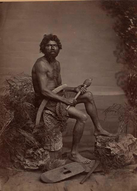 Studio-staged scene of an Aboriginal man of New South Wales, sitting and holding a club