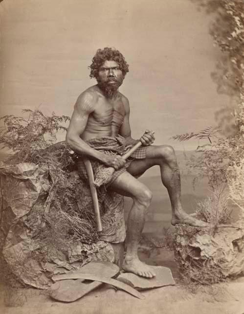 Studio-staged bush scene featuring an Aboriginal man of New South Wales, seated and holding a club