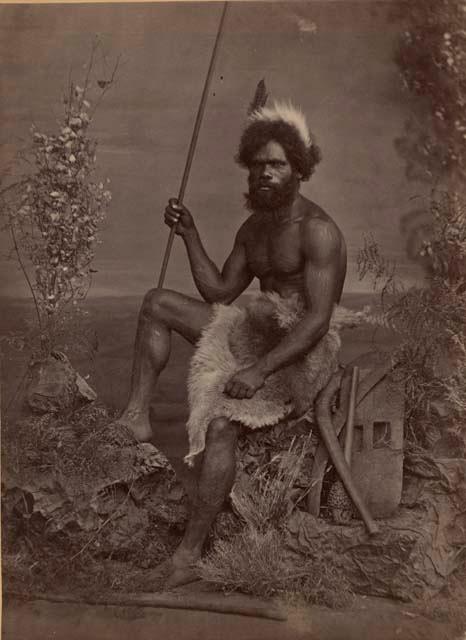 Studio-staged bush scene featuring an Aboriginal man of New South Wales, seated and holding a spear