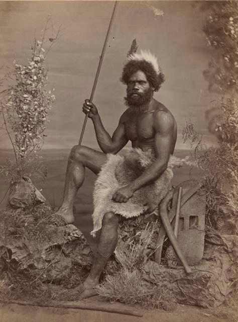 Studio-staged bush scene featuring an Aboriginal man of New South Wales, seated and holding a spear