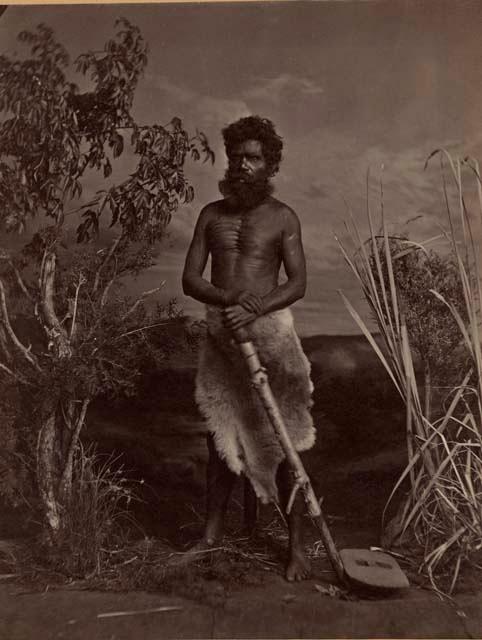 Studio-staged bush scene featuring an Aboriginal man of New South Wales, holding a thick branch