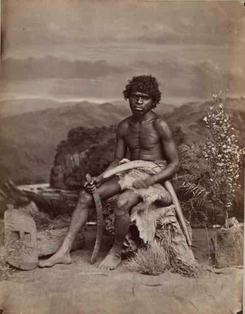 Studio-staged bush scene featuring an Aboriginal man seated and holding two boomerangs