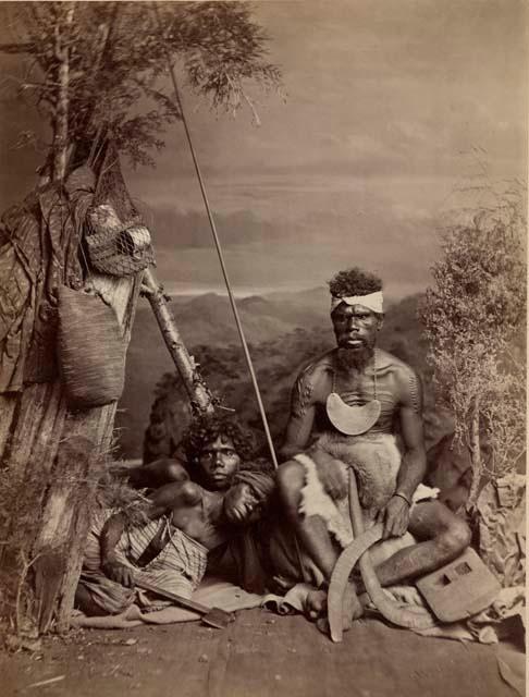 Studio-staged bush scene featuring three Aboriginal adults, a wooden shelter, boomerangs, and an axe