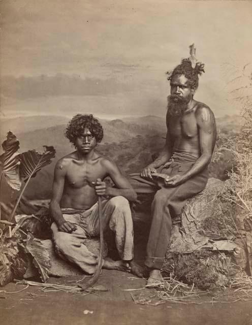 Studio-staged bush scene featuring two Aboriginal men seated and holding boomerangs