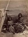 Studio-staged bush scene featuring two Aboriginal women and a child seated near a wooden shelter