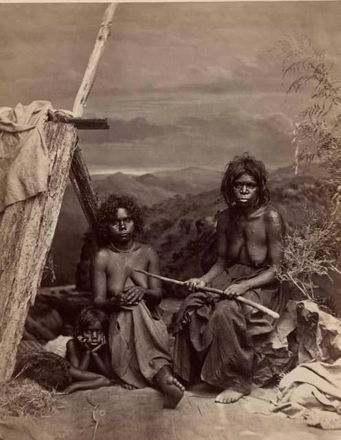 Studio-staged scene featuring two Aboriginal women and a child sitting near a wooden shelter