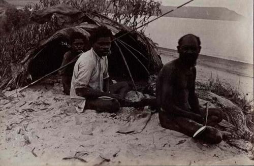 Two Aboriginal men and one young boy seated on sand before a small shelter