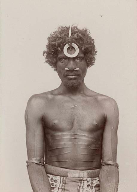 Portrait of Aboriginal Man wearing bone through septum and decorated headpiece, ritual scarification on abdomen.