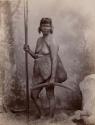 Studio shot of Aboriginal Woman wearing Fur Pelt, holding Two Spears and Two Boomerangs and carrying a Net Bag