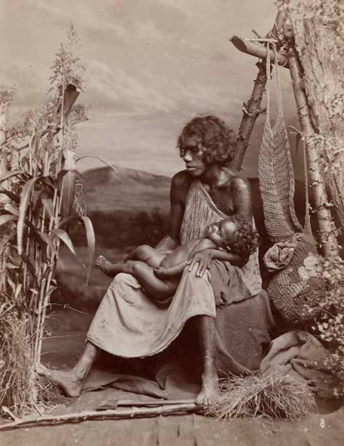 Studio-stage Bush scene showing Aboriginal Woman holding Child, Net Bags hanging behind them