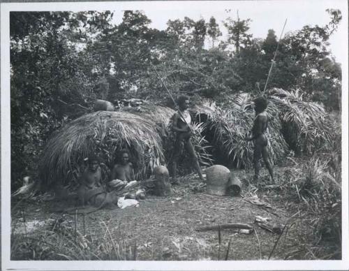 Two Aboriginal women sitting in front of shelters in a clearing, with two Aboriginal men standing and holding spears near them