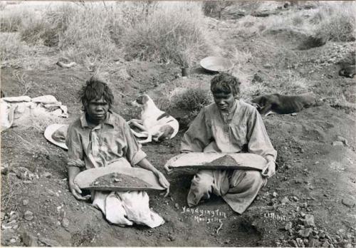 Two Aboriginal women sitting on ground in scrubland holding coolamons filled with dirt on their laps, dogs lying behind them