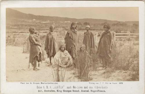 Seven female aboriginals draped in cloaks, two women sit on the ground and five stand behind them.