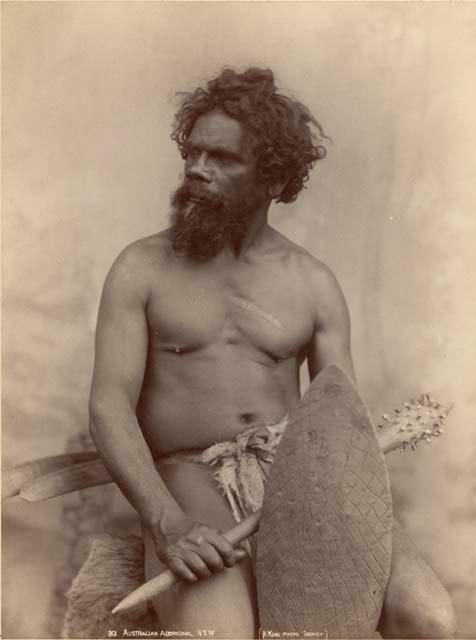 Studio portrait of Aboriginal man seated, holding a studded wooden club and a shield