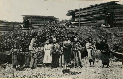 Five men, three women, four children, two dogs standing with wooden barrack-like