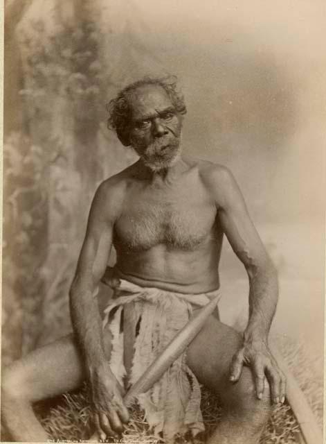 Studio staged portrait of an Aboriginal man, he is seated and holds a club