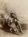Studio-staged Bush scene with an Aboriginal woman sitting, holding a boomerang and wearing a net bag