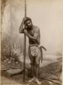 Studio-staged bush scene featuring an Aboriginal man wearing an animal waist cover with two boomerangs tucked in, leaning on two wooden spears