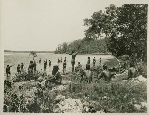 Large group of Aboriginal men near a river, some fishing others sitting and standing on shore