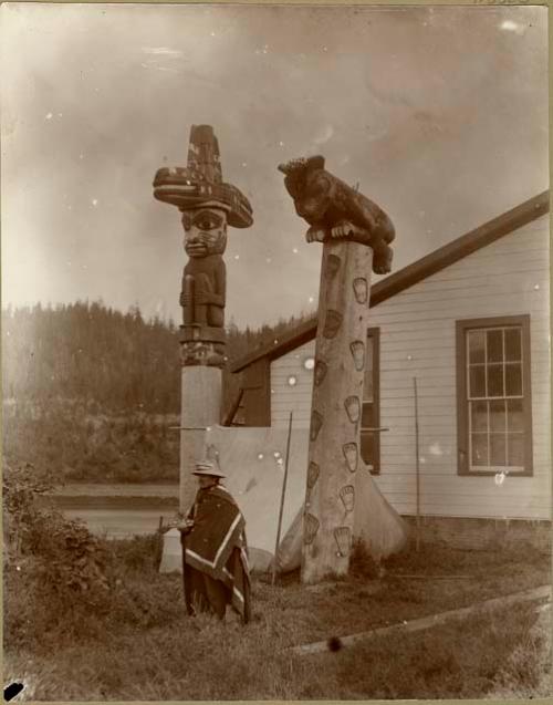 Chief Shakes standing before his house and totem poles