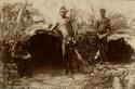 Photo of three Aboriginal men at two shelters, two men are holding spears, man in center holds a pipe in his mouth