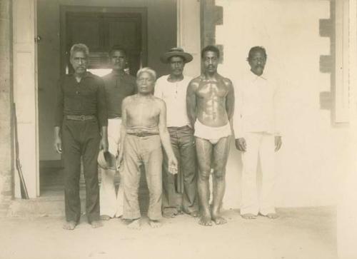 Group of sIx men stand by a doorway-- four are fully clothed, one is shirtless showing tattoos, and one has his full torso (oiled) and legs (tattooed) exposed.